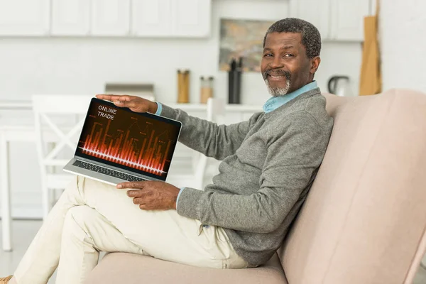 Uomo afroamericano sorridente seduto sul divano e mostrando laptop con app commerciale online sullo schermo — Foto stock