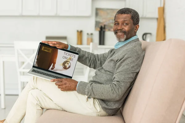 Sorrindo homem americano africano sentado no sofá e mostrando laptop com bilhetes aplicativo on-line na tela — Fotografia de Stock