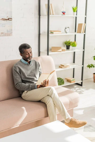 Senior african american man sitting on sofa in living room and reading book — Stock Photo