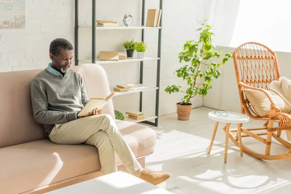 Senior african american man sitting on sofa in living room and reading book — Stock Photo