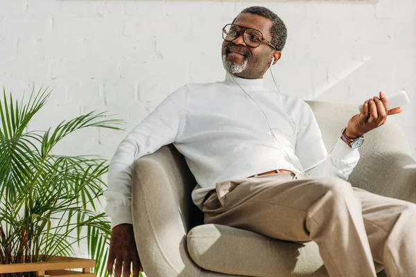 Élégant homme afro-américain assis dans un fauteuil et écouter de la musique dans les écouteurs — Photo de stock