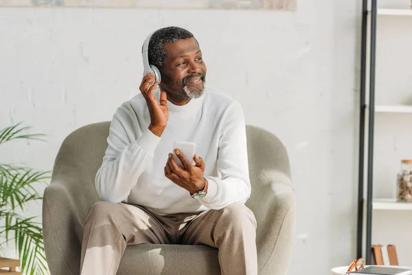 Hombre afroamericano elegante sentado en sillón y escuchando música en auriculares - foto de stock