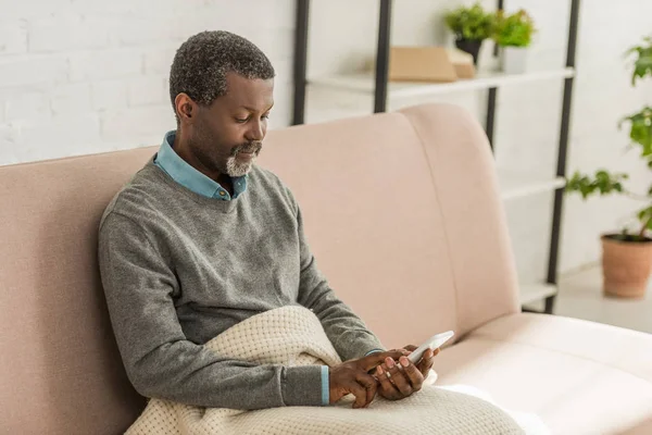 Thoughtful african american man sitting on sofa and using smartphone — Stock Photo