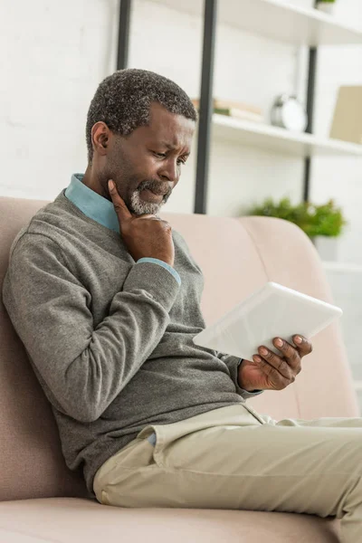 Thoughtful african american man sitting on sofa and using digital tablet — Stock Photo