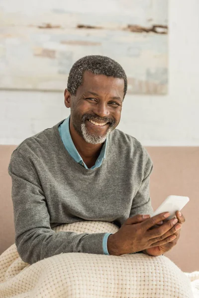 Cheerful african american man sitting with blanket on knees, holding smartphone and smiling at camera — Stock Photo