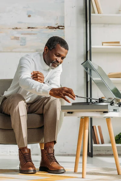 Stilvoller afrikanisch-amerikanischer Mann sitzt im Sessel und hört Musik auf Plattenspieler — Stockfoto