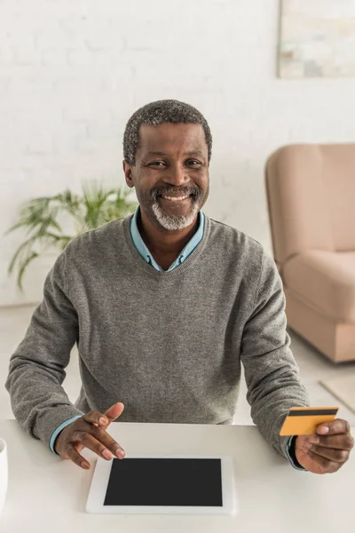 Alegre afroamericano sonriendo a la cámara mientras sostiene la tarjeta de crédito y el uso de la computadora portátil - foto de stock