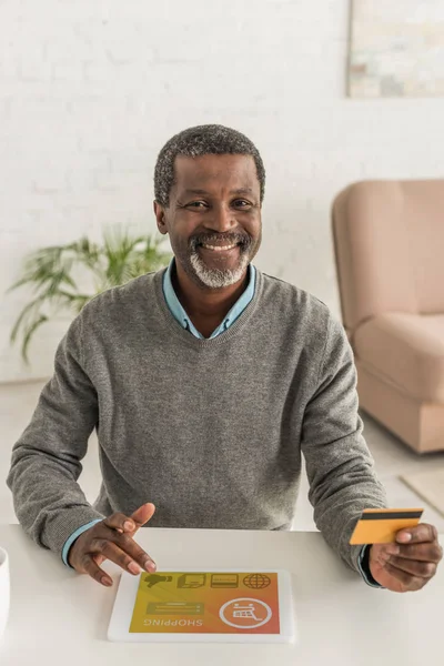 Hombre afroamericano alegre celebración de la tarjeta de crédito y el uso de tableta digital con aplicación de compras en línea en la pantalla - foto de stock