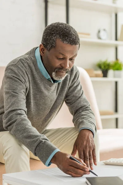 Aufmerksamer afrikanisch-amerikanischer Mann schreibt Stromrechnung, während er auf dem Sofa sitzt — Stockfoto