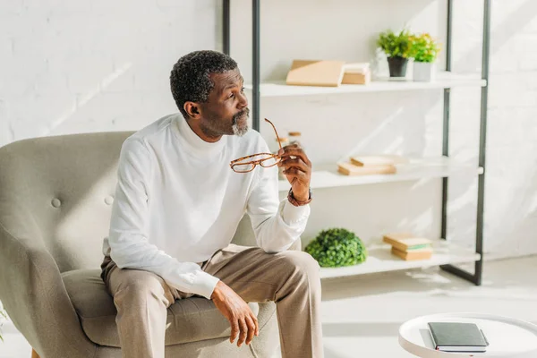 Sognante uomo africano americano seduto in poltrona, tenendo gli occhiali e guardando altrove — Foto stock