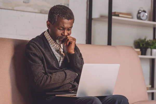 Hombre afroamericano molesto mirando el ordenador portátil mientras está sentado en el sofá - foto de stock