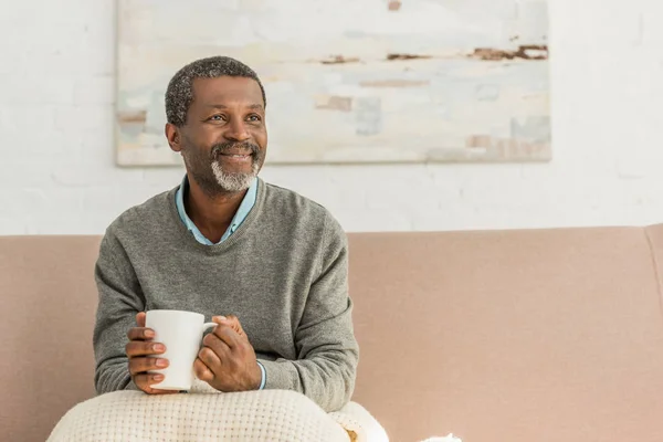 Sénior, hombre afroamericano positivo sentado con manta en las rodillas y sosteniendo taza de bebida caliente - foto de stock