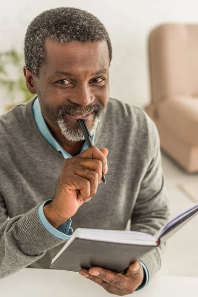 Homem americano africano alegre segurando caneta e caderno enquanto sorrindo para a câmera — Fotografia de Stock