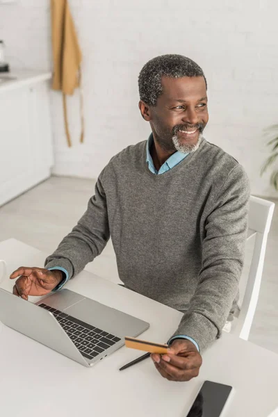 Fröhlicher afrikanisch-amerikanischer Mann schaut weg, während er neben Laptop sitzt und Kreditkarte hält — Stockfoto