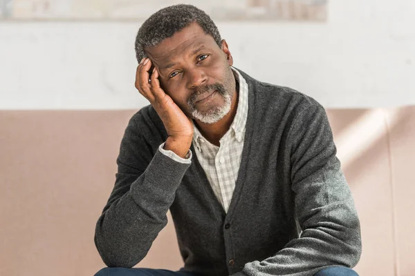 Rêveur afro-américain assis sur le canapé à la maison et regardant la caméra — Photo de stock