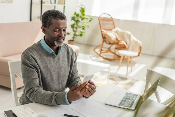 Ernstzunehmender afrikanisch-amerikanischer Mann schaut auf smartpone, während er mit Versorgungsrechnungen am Tisch sitzt — Stockfoto