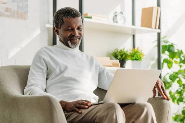 Smiling african american man sitting in armchair and using laptop — Stock Photo
