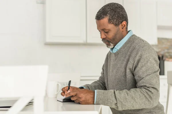 Hombre afroamericano serio sentado a la mesa y escribiendo en un cuaderno - foto de stock