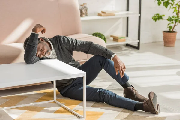 Deprimido afro-americano homem inclinado na mesa enquanto sentado no chão em casa — Fotografia de Stock