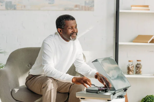 Happy african american man sitting in armchair and listening music on record player — Stock Photo