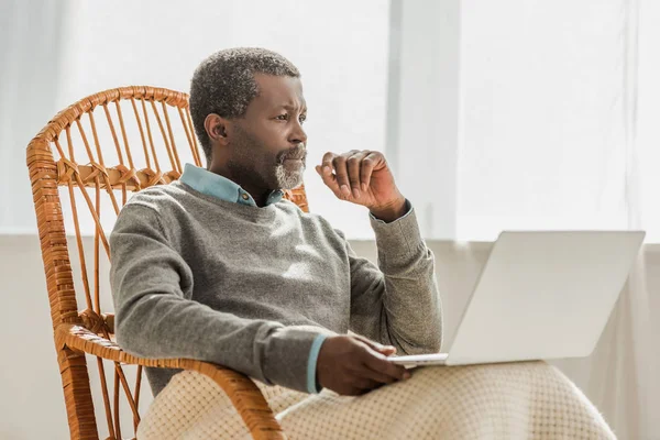 Hombre afroamericano soñador sentado en silla de mimbre con portátil y mirando hacia otro lado - foto de stock