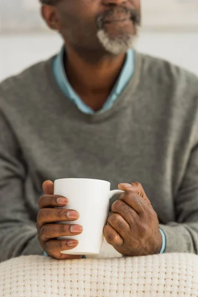 Vista cortada do homem americano africano sênior sentado com cobertor nos joelhos e segurando copo com bebida de aquecimento — Fotografia de Stock