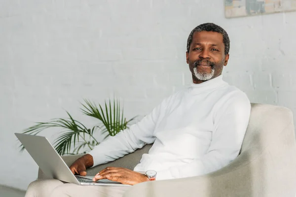 Senior homme afro-américain assis dans un fauteuil avec ordinateur portable et souriant à la caméra — Photo de stock