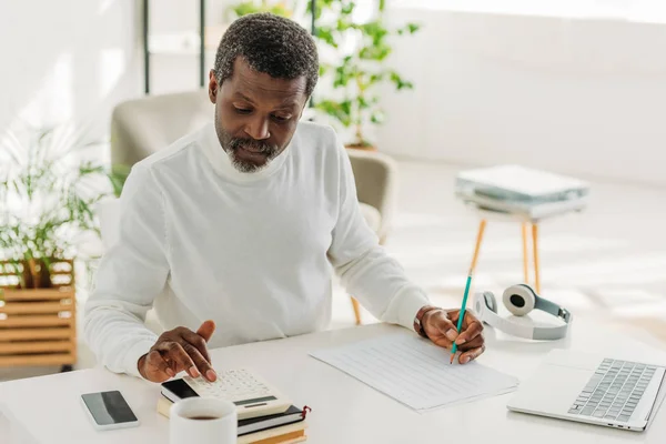 Serious african american man sitting at table near utility bill and calculating expenses — Stock Photo