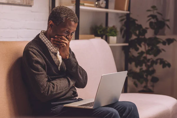 Chateado afro-americano homem sentado no sofá e olhando para laptop — Fotografia de Stock