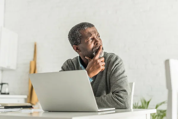 Pensativo homem afro-americano olhando para longe enquanto sentado perto do laptop — Fotografia de Stock