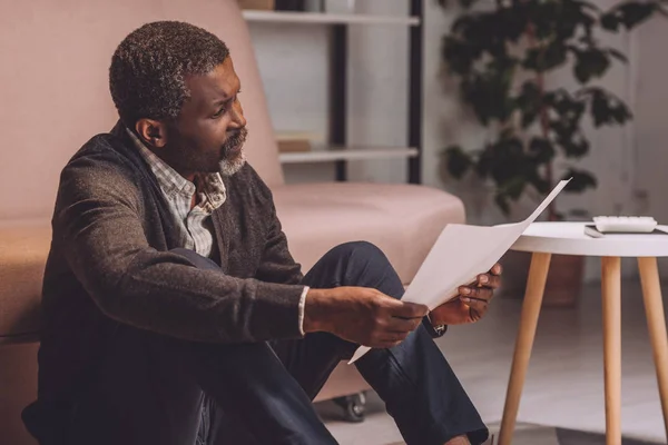 Upset african american man sitting on floor and looking at utility bill — Stock Photo