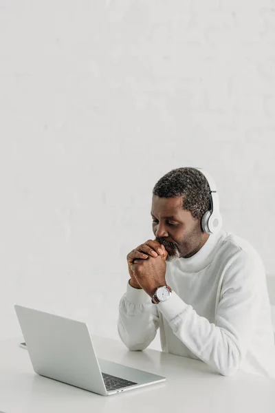 Hombre afroamericano serio en auriculares sentados cerca de la computadora portátil - foto de stock