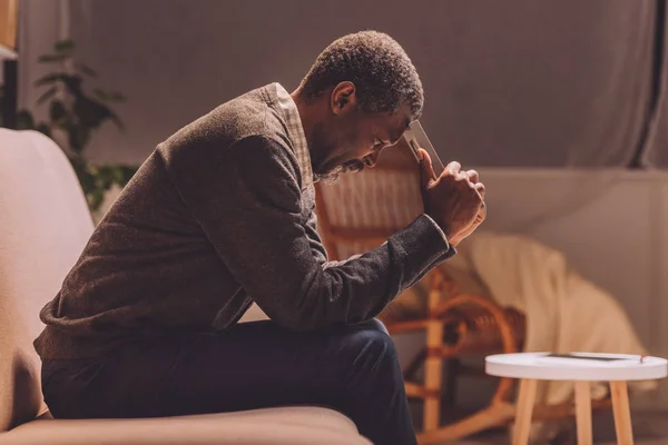 Depressed african american man sitting on sofa at night and holding photo frame near head — Stock Photo