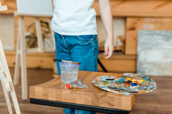 Cropped view of kid standing near wooden table with gouache paints — Stock Photo