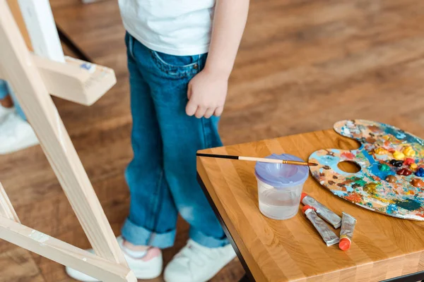 Vue recadrée de l'enfant debout près de la table en bois avec des peintures gouache — Photo de stock