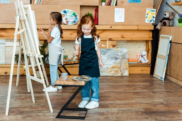 Cute redhead kid holding paintbrush near colorful gouache paints and child — Stock Photo