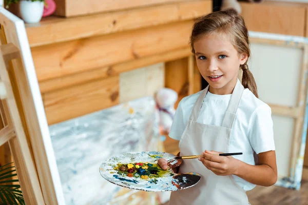 Happy kid in apron holding palette with colorful gouache paint — Stock Photo