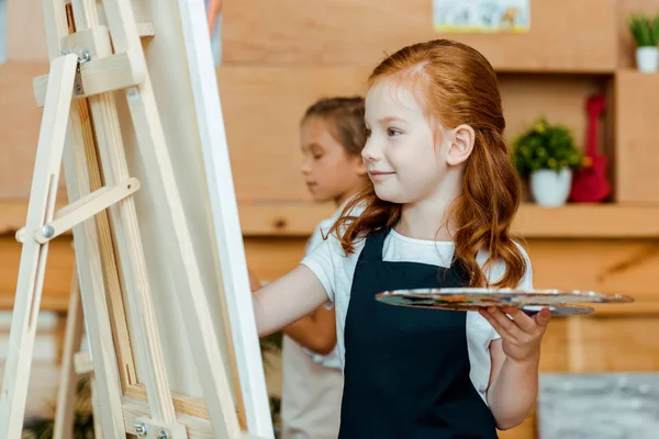 Fuoco selettivo di bambino rosso allegro che tiene la tavolozza vicino a cavalletto e bambino in scuola d'arte — Foto stock