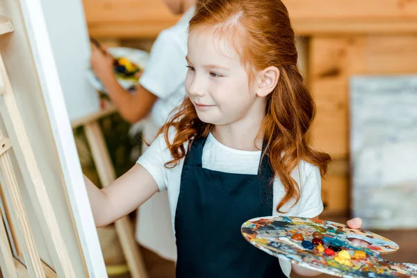 Messa a fuoco selettiva di allegra rossa bambino tenendo tavolozza colorata vicino cavalletto e bambino nella scuola d'arte — Stock Photo