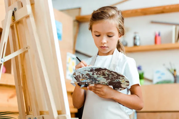 Selective focus of kid in apron holding palette with colorful gouache paint — Stock Photo