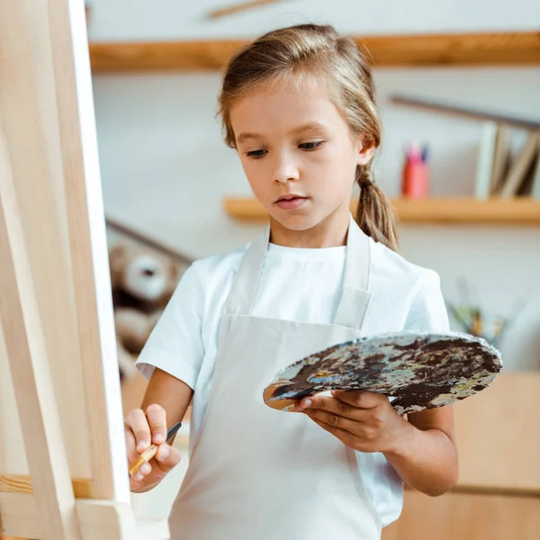 Selective focus of kid in apron holding palette with colorful gouache paint and paintbrush — Stock Photo