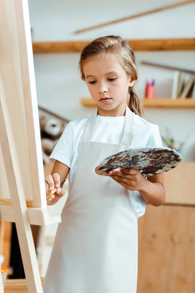 Kid in apron holding palette with colorful gouache paint and paintbrush — Stock Photo