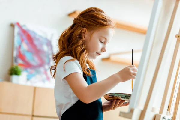 Messa a fuoco selettiva di cute rossa bambino in possesso di tavolozza e pennello vicino cavalletto — Stock Photo