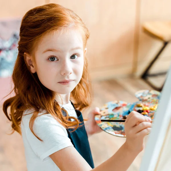 Selective focus of cute redhead child holding paintbrush and looking at camera — Stock Photo
