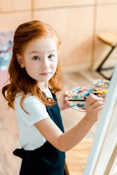 Selective focus of redhead child holding paintbrush and looking at camera — Stock Photo