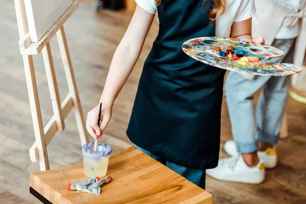 Ausgeschnittene Ansicht eines Kindes mit Palette und Pinsel in der Nähe eines Kindes in der Kunstschule — Stockfoto