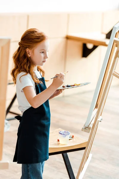 Cute redhead kid with paintbrush and palette near easel — Stock Photo