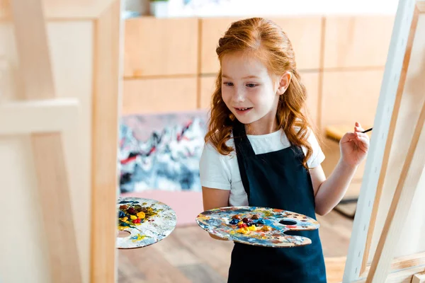 Selective focus of redhead kid looking at easel while holding palette — Stock Photo