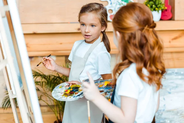 Селективное внимание милого ребенка, смотрящего на мольберта рядом с рыжим ребенком в художественной школе — стоковое фото