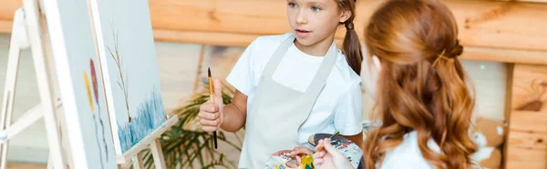 Panoramic shot of cute kid looking at easel with painting near redhead child in art school — Stock Photo
